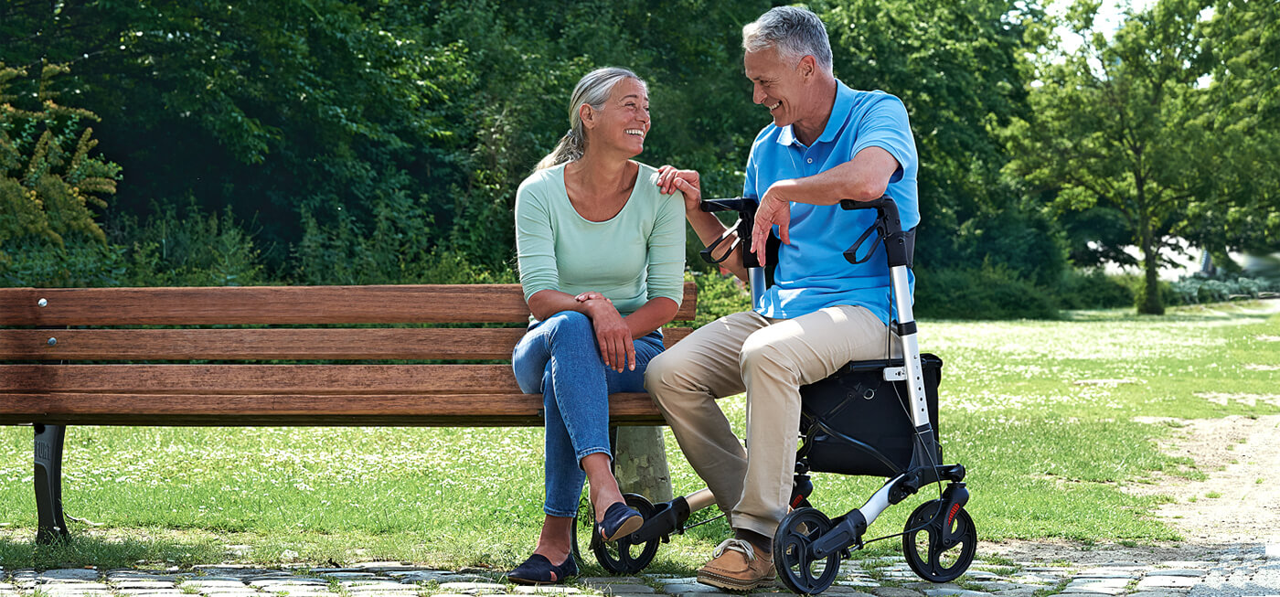 Eine Frau sitzt auf einer Parkbank, daneben ihr Mann, der auf einem Rollator sitzt.