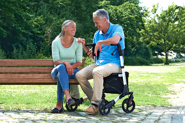 Eine Frau sitzt auf einer Parkbank, daneben ihr Mann, der auf einem Rollator sitzt.